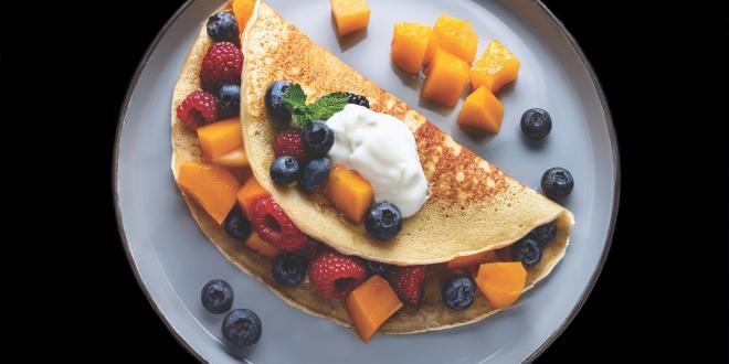 A pancake wrapped around cantaloupe chunks, raspberries, and blueberries, with a dollop of yogurt on top.