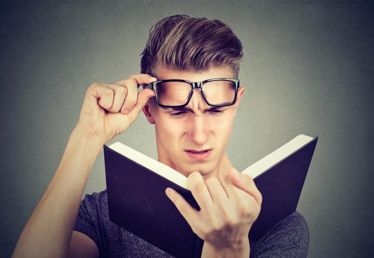 A young man lifting his glasses straining to read a book.